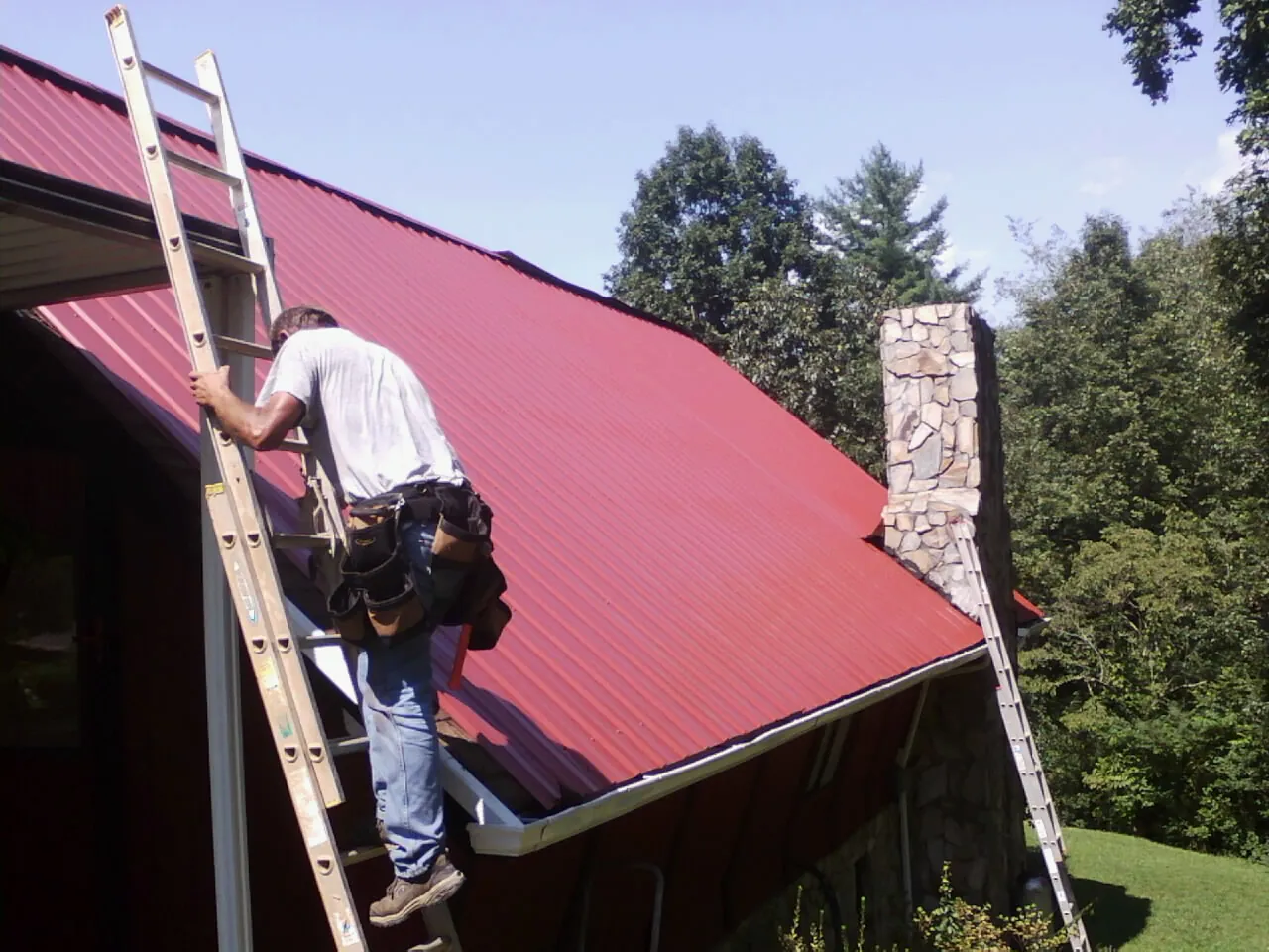 Roofer on a ladder beside a red metal roof and stone chimney — Soffit & Fascia Repair by Roofing Brothers in Overbrook